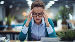 Stressed female office employee holding her head in frustration at a desk filled with paperwork, illustrating workplace burnout and the quiet cost of overtime.