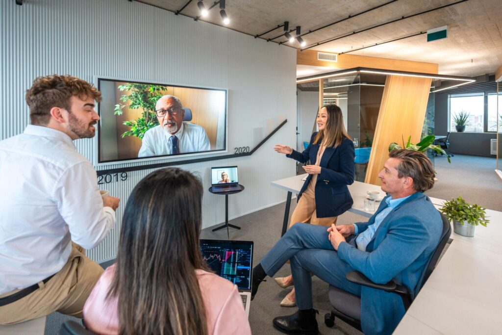 A hybrid team collaborates during a meeting, with employees working in the office while remote colleagues participate through a video conference, illustrating today's flexible workplace.