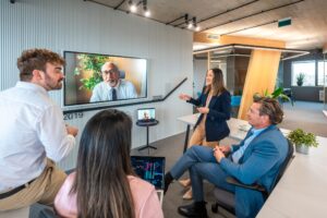 A hybrid team collaborates during a meeting, with employees working in the office while remote colleagues participate through a video conference, illustrating today's flexible workplace.